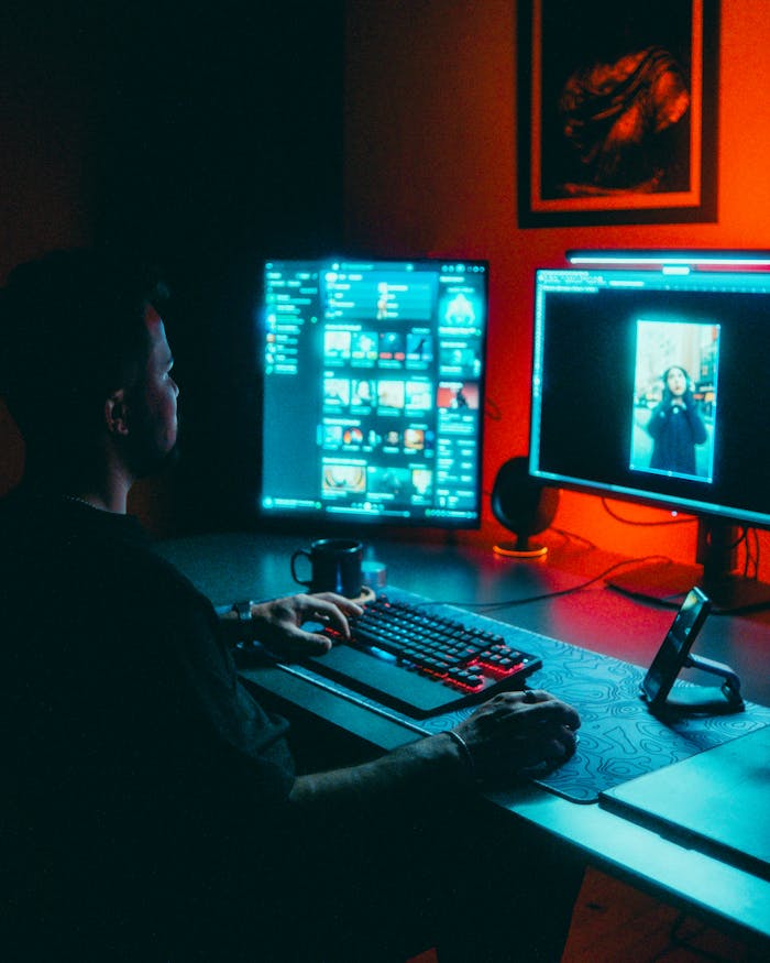 A man working on dual monitors in a dark room with red ambient lighting.